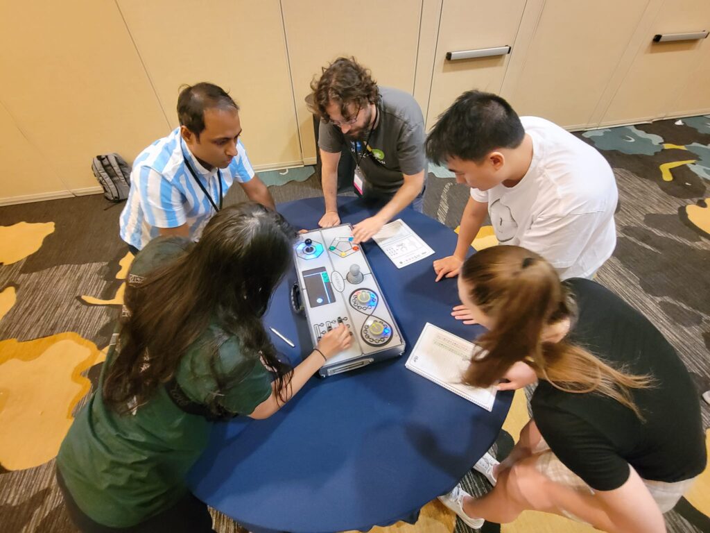 Five participants leaning over a Team Synergy retro gaming console, strategizing and solving mini-game challenges during a team building activity.