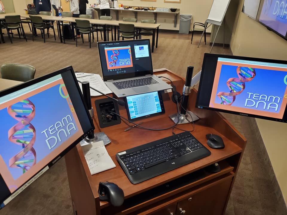 A workstation set up for the Team DNA session, featuring multiple screens displaying the event's branding and presentation materials.