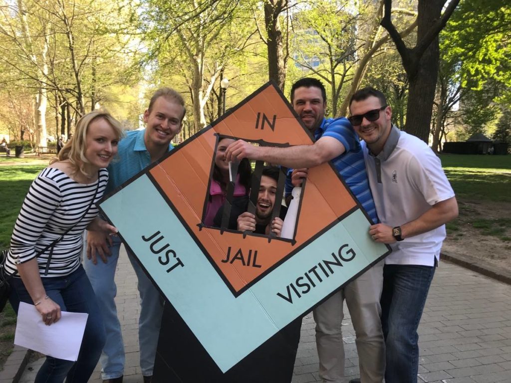 A group of six people smiling and posing with a large board game prop during a Team-opoly scavenger hunt, with one person playfully positioned behind the 'In Jail' section of the board.