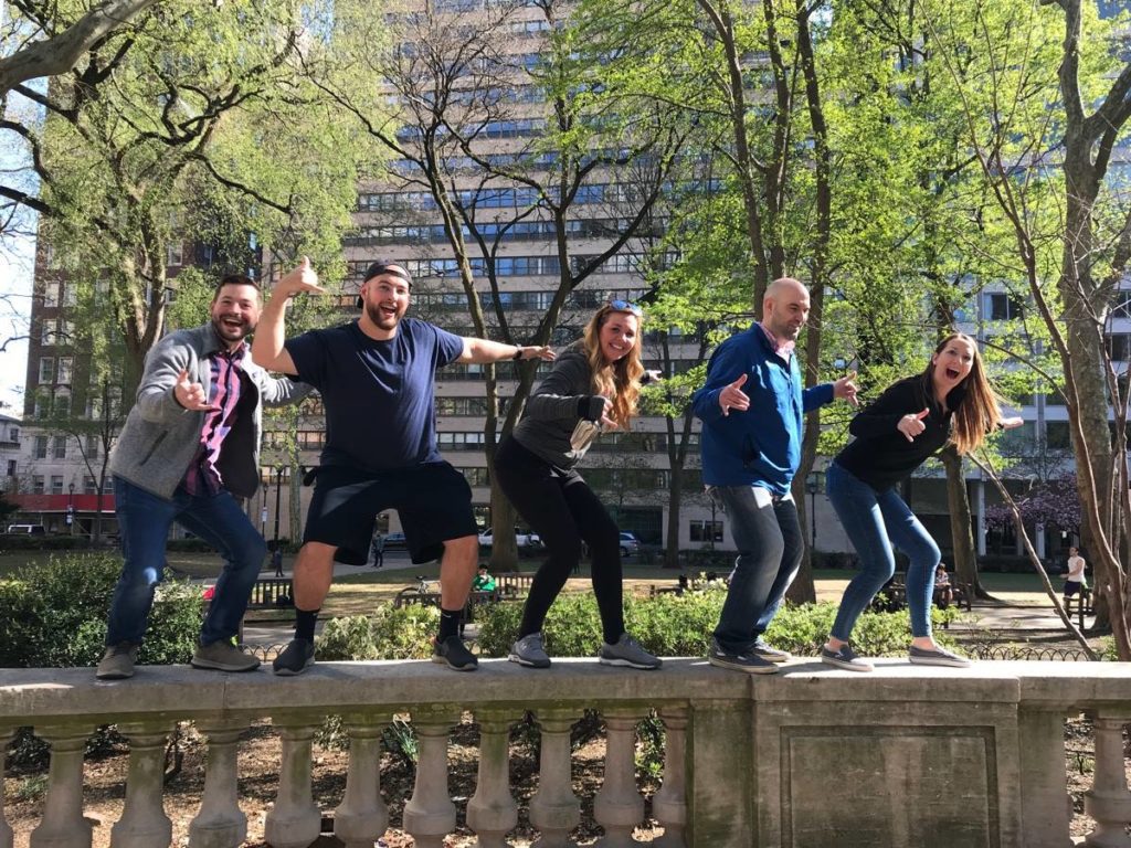 A team of five participants standing on a low stone wall, posing playfully during a Team-opoly scavenger hunt in a city park.