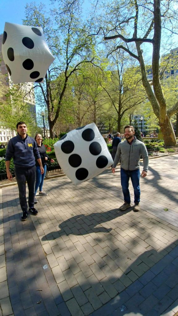 Participants tossing large, oversized dice during a Team-opoly scavenger hunt in a city park, surrounded by trees and urban buildings.