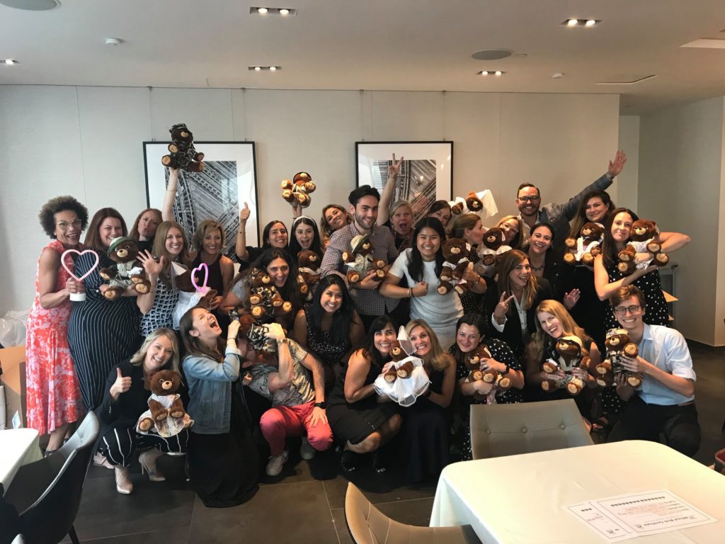 A large group of enthusiastic participants, smiling and posing with teddy bears they have assembled during a team building event. The group, made up of men and women, holds up their brown teddy bears. Many are raising their hands or posing playfully, creating a joyful and celebratory atmosphere.