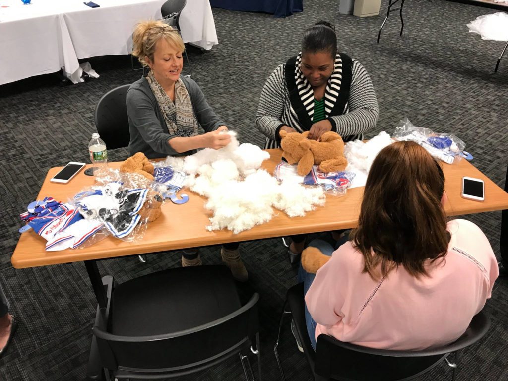 Three women seated at a table during a teddy bear assembly team building event. They are focused on stuffing and creating teddy bears from kits that include plush materials, stuffing, and small accessories. Various bear parts and materials are scattered on the table, and they appear to be enjoying the hands-on activity.