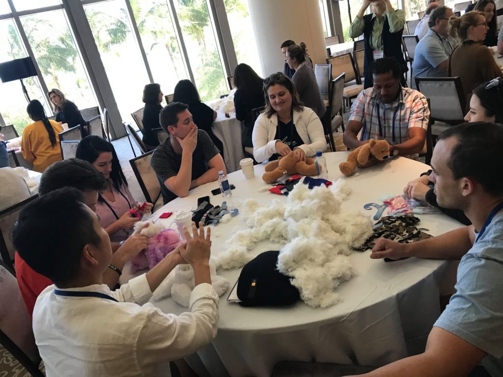 A group of individuals sits around a table working on a Team Teddy Rescue Bear event. Participants are stuffing teddy bears and organizing materials like cotton filling, bear parts, and accessories. The room is filled with other groups, all engaged in similar activities.