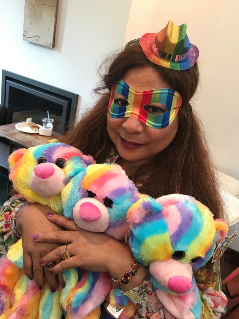 A woman dressed festively in a rainbow-colored mask and a mini hat, holding three brightly colored rainbow teddy bears. She has a warm expression as she hugs the bears. The setting appears to be a fun and playful environment, part of a charitable team building event.