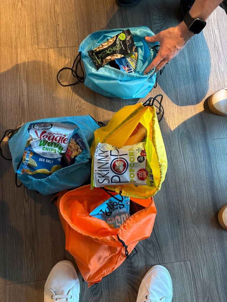 Colorful drawstring bags filled with healthy snacks such as veggie chips, pistachios, and popcorn, ready to be donated as part of the Healthcare Heroes scavenger hunt team building program.