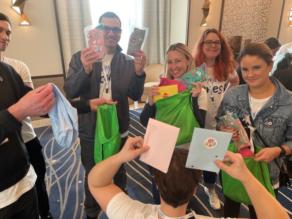 A group of smiling participants holds up cards and green bags filled with items like socks and snacks. They are engaged in assembling appreciation bags for healthcare professionals during a Healthcare Heroes scavenger hunt team building activity.