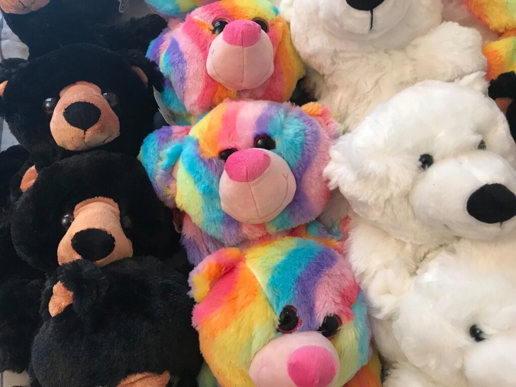 A close-up view of plush teddy bears, showing a variety of colors and designs. Some bears are black with brown accents, while others are brightly colored in a rainbow pattern with pink noses. There are also white polar bear plushies with black noses. The bears are neatly arranged, creating a cheerful, soft display, ready to be customized.