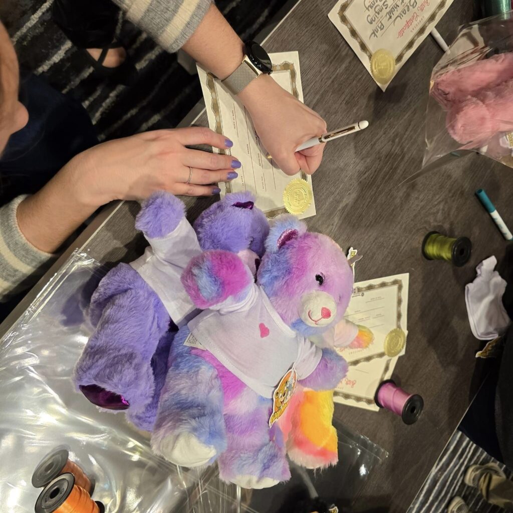 Person writing a certificate next to colorful stuffed bears during the Team Teddy Bear Rescue activity at a corporate charitable team building event.