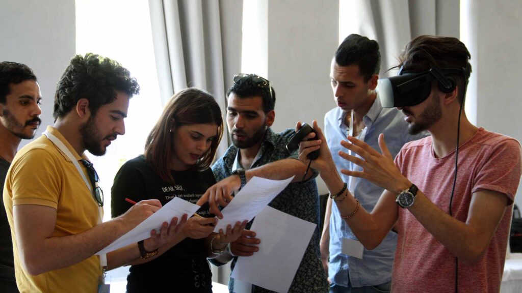 A group of participants collaborates on a task, with one member wearing a VR headset and explaining the virtual challenge to the rest of the team during The Infinite Loop team building event.