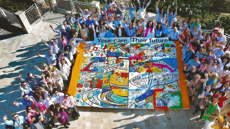 A large group of participants gathers around a completed mural during The Big Picture team building event, celebrating their collaborative artwork with hands raised in excitement. The vibrant mural emphasizes teamwork and shared goals.