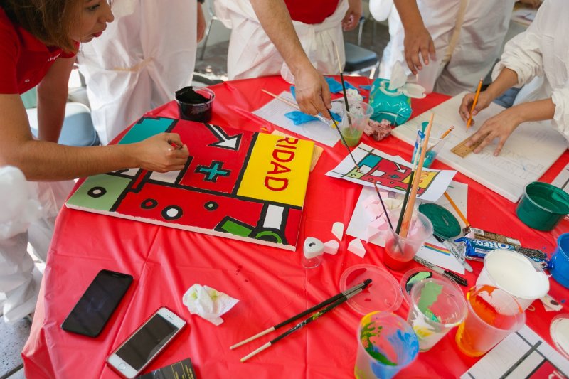 Participants work together on a colorful mural panel during The Big Picture team building event. The table is filled with paint supplies, and one of the participants carefully paints a section featuring bold shapes and colors.