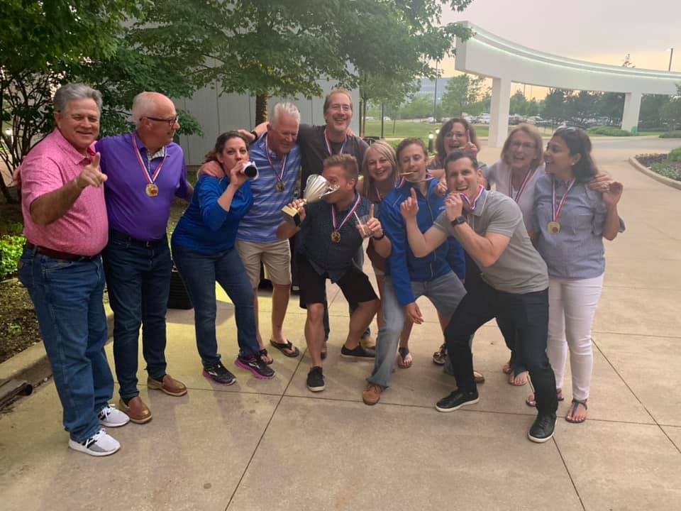 A group of smiling adults posing together outdoors, celebrating their victory at a team building event. They are wearing medals around their necks and holding a trophy, displaying a joyful and triumphant spirit. Some team members are playfully gesturing or mimicking drinking from the trophy, showcasing their camaraderie and enthusiasm.