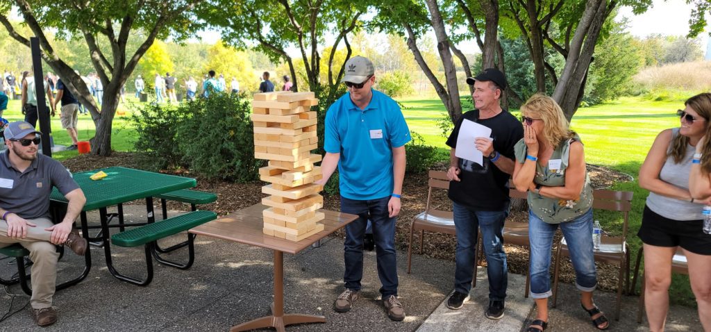 The image shows a group of participants engaged in a giant Jenga game. The participants are focused on the game, watching closely as one person carefully removes a block from the tall, precariously stacked tower.