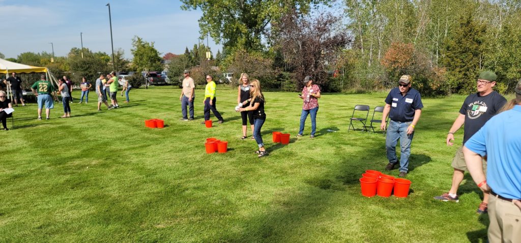 Adults participating in an outdoor team building activity on a sunny day. They are standing on a grassy field, spaced apart, taking part in the Ultimate Tailgate Challenge.