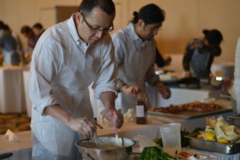 A participant at the 'Food Truck Challenge' is preparing a dish. Wearing an apron and gloves, he focuses on mixing ingredients in a large bowl. Behind him, other participants are also engaged in their culinary tasks, surrounded by fresh ingredients