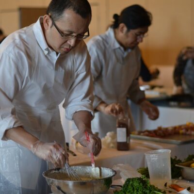 A participant at the 'Food Truck Challenge' is preparing a dish. Wearing an apron and gloves, he focuses on mixing ingredients in a large bowl. Behind him, other participants are also engaged in their culinary tasks, surrounded by fresh ingredients