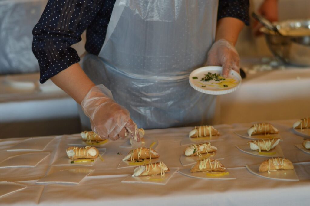 The image shows a participant carefully preparing and plating small tacos as part of a team building food truck challenge. The individual uses gloves for food safety while arranging the tacos neatly on a table covered with a white cloth. Each taco is garnished with a drizzle of sauce, and the participant holds a small plate with additional garnishes, ready to add the finishing touches.