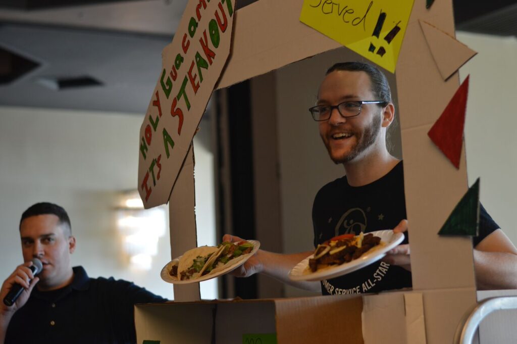 Participants enthusiastically showcase their creative food truck setup during a 'Food Truck Challenge' team building event. A team member stands under a handcrafted food truck façade, proudly presenting plates of tacos.