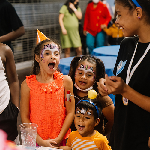 Three children with painted faces are excitedly smiling and laughing at a party. The child in the front is wearing an orange party hat, and they are surrounded by other attendees and decorations, creating a festive atmosphere.