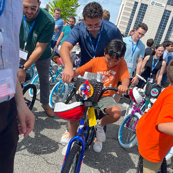 A group of volunteers assisting children in riding their newly assembled bikes during a Charity Bike Build event. A boy in an orange shirt excitedly rides a decorated blue bike, guided by a volunteer.