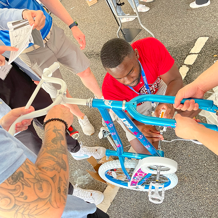 A group of participants working together to assemble a children's bicycle during a Charity Bike Build event. One person is tightening a bolt while others hold the bike steady and refer to instructions, demonstrating teamwork and collaboration.