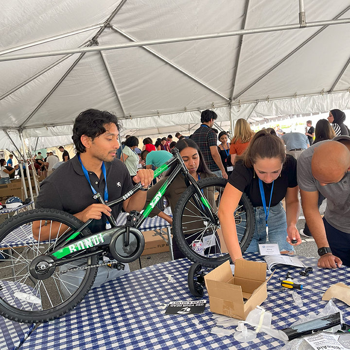 A group of participants working together to assemble a green bicycle during a Charity Bike Build event. They are under a large tent, focused on attaching parts with tools. Other groups in the background are also working on assembling bikes.