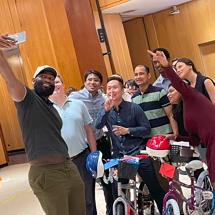 Group of enthusiastic participants taking a selfie during a Charity Bike Build event. They are smiling and posing with peace signs while standing next to the bicycles they assembled, which are adorned with helmets and decorations, highlighting the fun and collaborative atmosphere of the event.