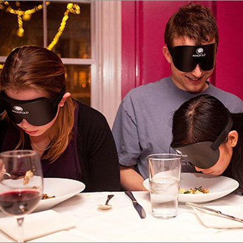 Three people wearing blindfolds while eating at a dining table, participating in a sensory deprivation dining experience to enhance communication and trust.