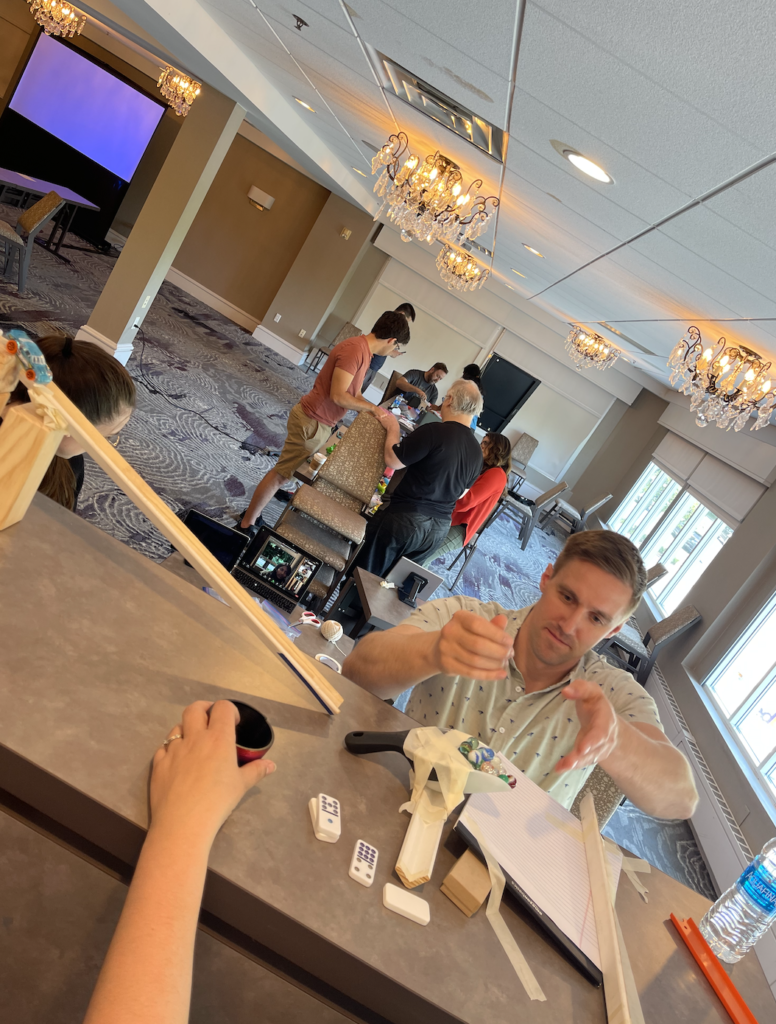 Participant carefully assembling a part of a contraption during the Domino Effect team building event, while other teams work in the background.