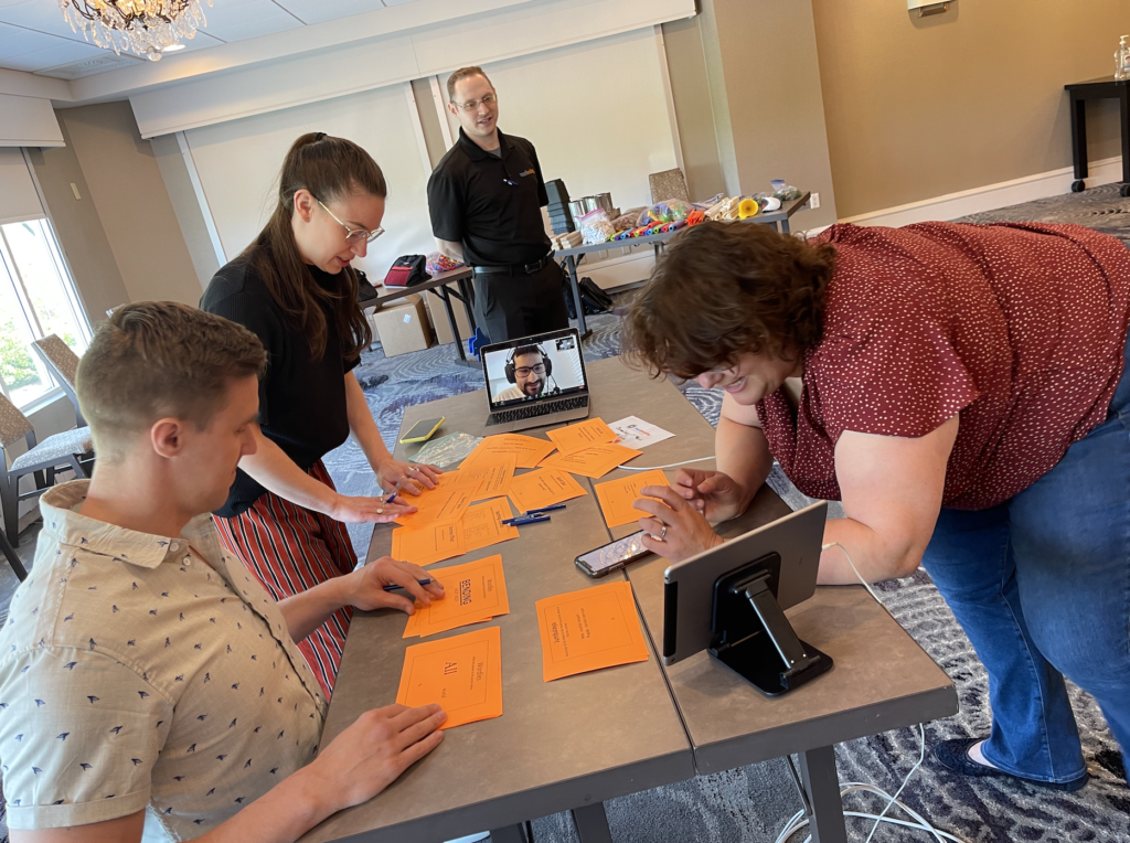 Team members collaborating over orange cards during the Domino Effect team building event, with a remote participant joining in via a tablet.