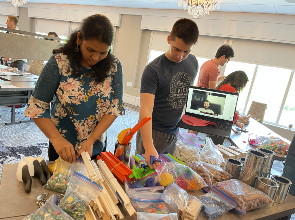 Participants sorting through materials and building supplies during the Domino Effect team building event, with a remote team member visible on a tablet.