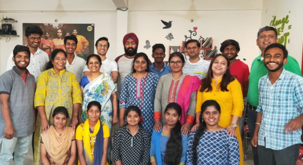 A group of men, women, and children from a rural community in India gather together for a group photo, smiling as they celebrate the donations they have received. The diverse group, dressed in colorful traditional and modern attire, reflects the joy and gratitude of the local community benefiting from the donation project.