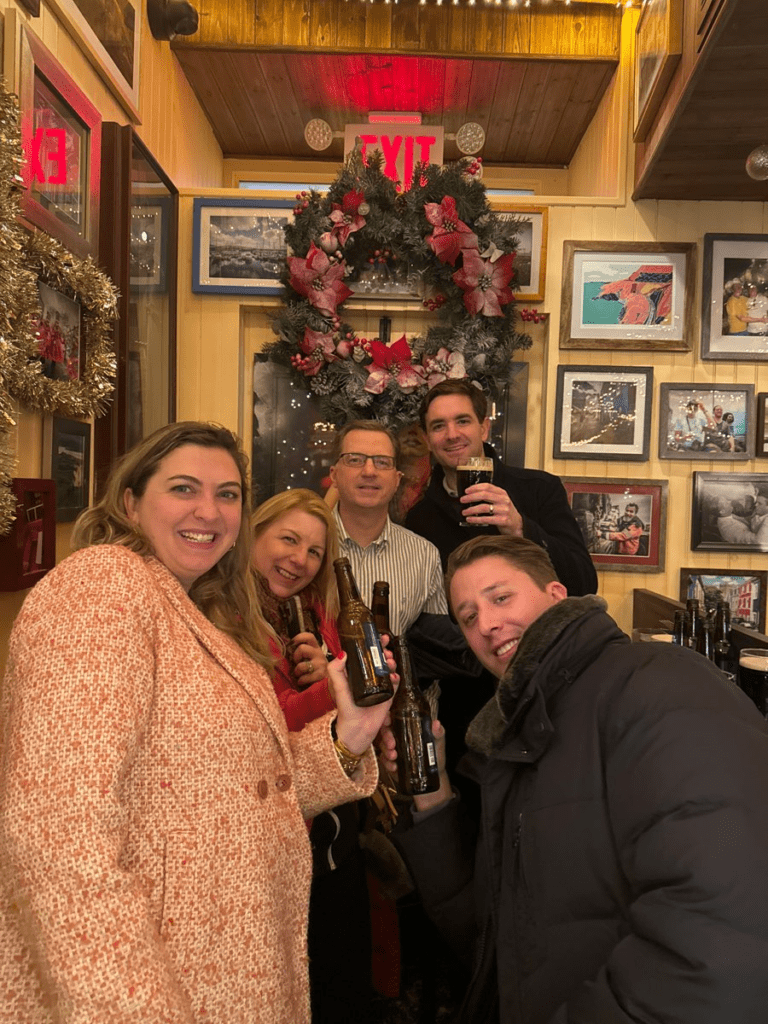 Smiling team members raise their drinks in celebration during a festive stop on the Pub Crawl Scavenger Hunt, enjoying the holiday decor and camaraderie.