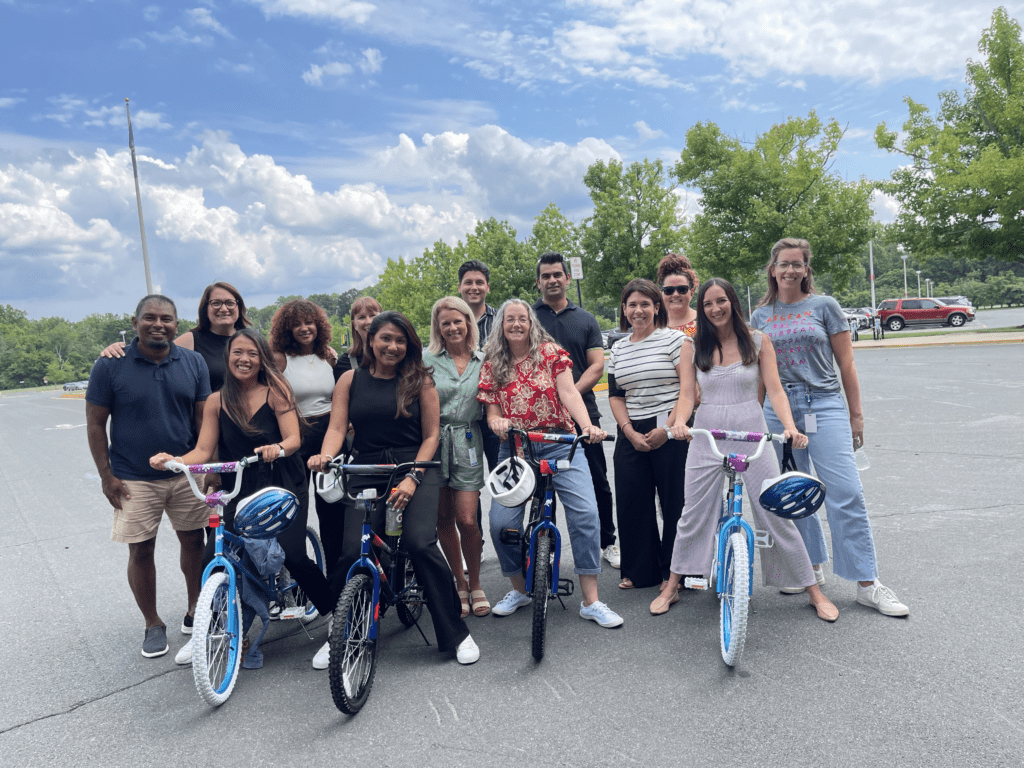 A group of smiling participants standing outdoors with two completed bicycles during a Charity Bike Build event. The team poses together in a parking, showing their assembled bikes with helmets on the handlebars.