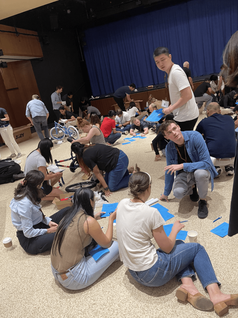 Participants gathered in a large room working in teams during a Charity Bike Build event. Some groups are sitting on the floor, writing on blue papers, while others are assembling bicycles. The scene is full of activity, with people collaborating and interacting, showcasing a productive and collaborative atmosphere.