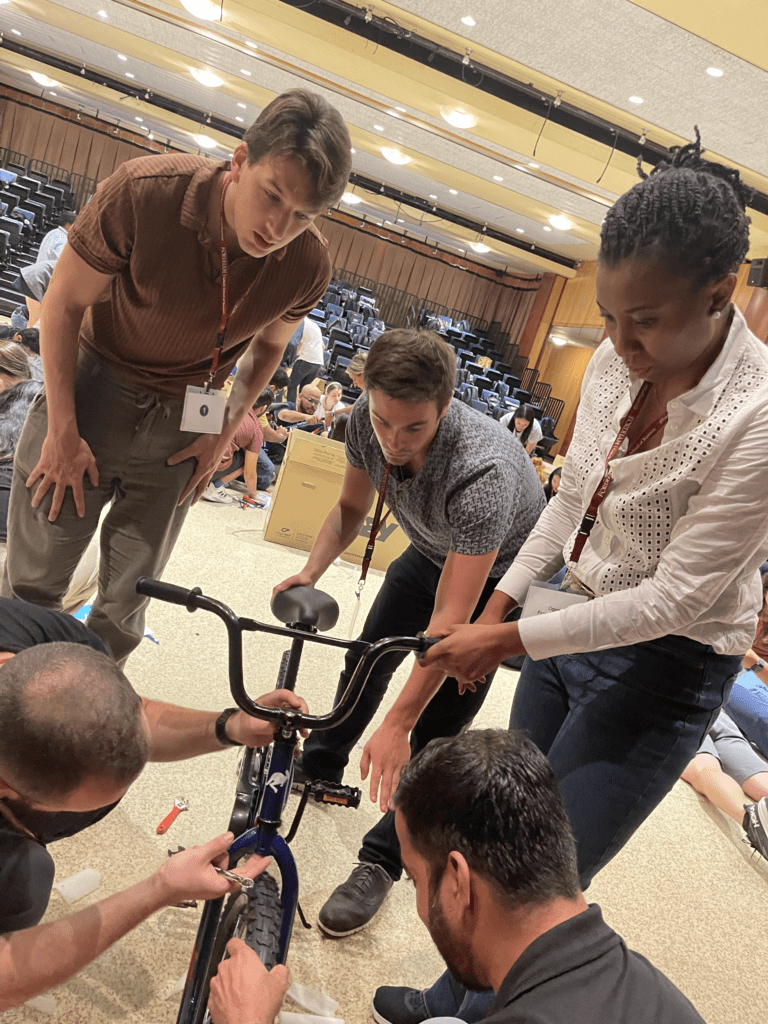 A group of four team members collaborate to assemble a bicycle during a Charity Bike Build event. Two participants hold the bike frame steady while another assists in attaching the wheels. The team works together in a large room filled with other participants working on similar tasks.