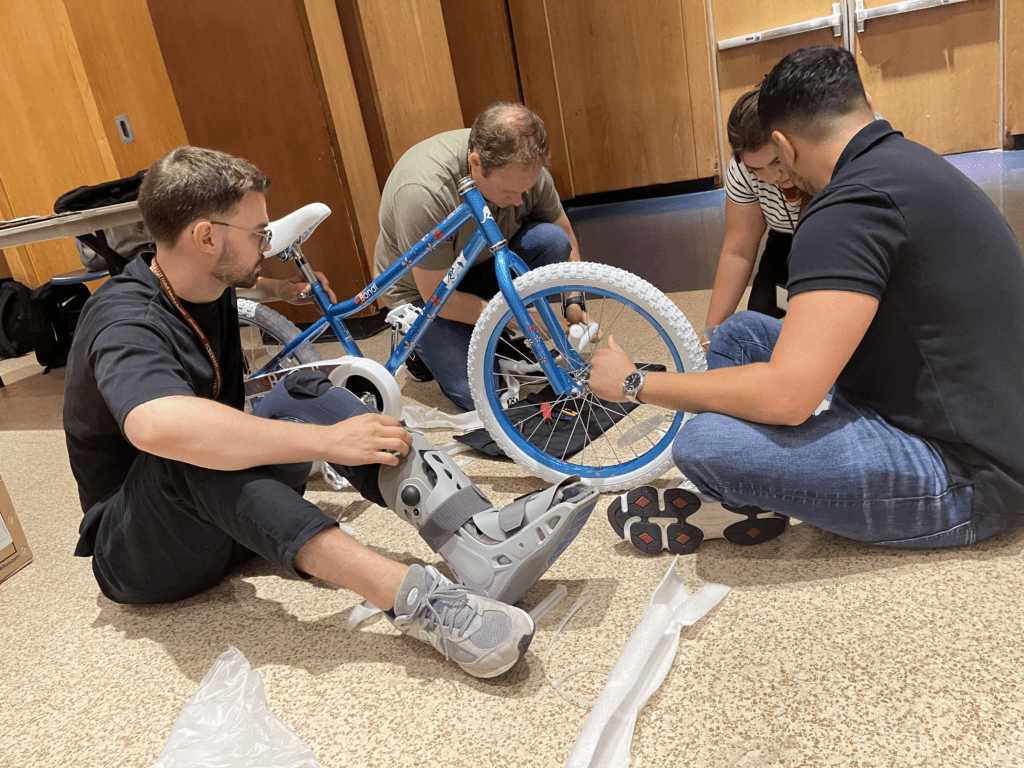 Group of four participants sitting on the floor working together to assemble a blue children's bike during a Charity Bike Build team building event. The participants are focused on attaching parts to the bike frame, demonstrating teamwork and collaboration.