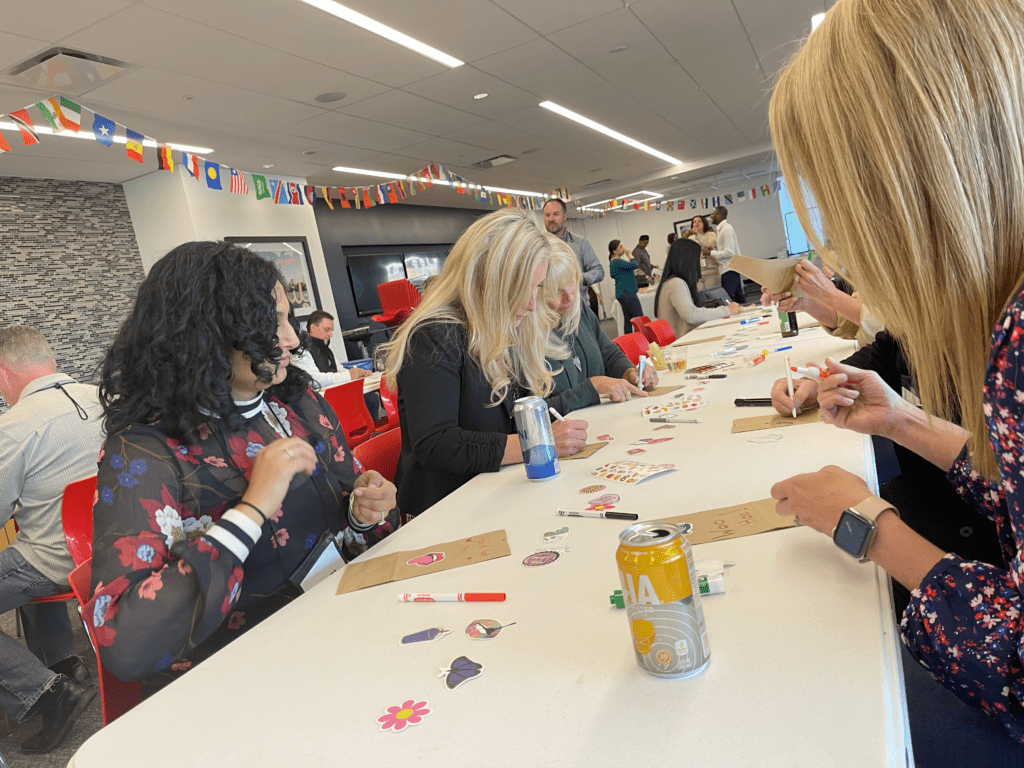 Participants sitting at a long table decorating brown paper bags with stickers and markers during The Great Snack Pack activity at a corporate charitable team building event.