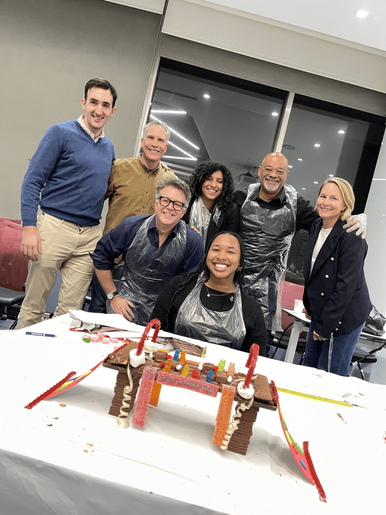 A team proudly poses with their creative, candy-decorated chocolate bridge after completing the Chocolate Challenge team building event, showcasing teamwork and fun.