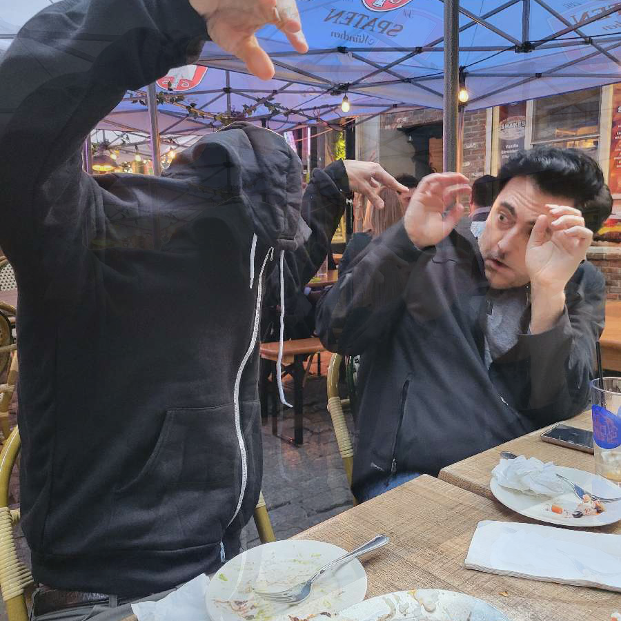 Two people sitting at an outdoor table appear to be acting out a playful, dramatic scene. The person on the left is wearing a hoodie with the hood pulled over their face, raising their arms as if to 'scare' the other person. The person on the right is recoiling in mock fear, raising their hands defensively while leaning back in their chair. The scene takes place under a canopy with string lights, and there are empty plates on the table, suggesting the duo is at a restaurant or cafe.