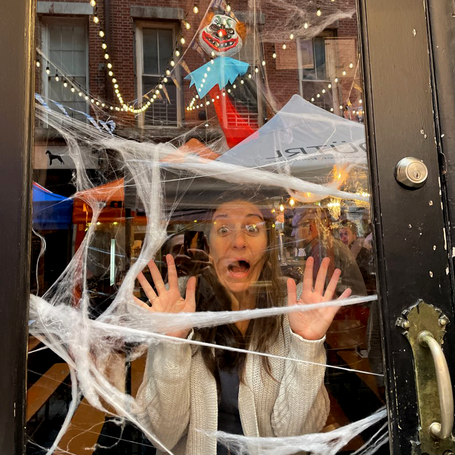 A woman stands behind a glass door decorated with fake spider webs, pressing her hands and face against the glass in a playful expression of fear, as though she's trapped. Behind her, festive lights are strung across an outdoor space, and a red-and-black decorative figure hangs above, adding to the Halloween atmosphere.