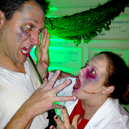 A man and a woman are dressed up in spooky, Halloween-themed makeup and costumes. The man has a startled, scared expression with his hands raised defensively, while the woman, who has bloody makeup with a gash across her face, is leaning toward him with her mouth open, mimicking a scream or growl.