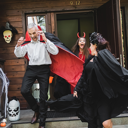 A group of three people dressed in Halloween costumes standing on a porch, engaging in playful action. The man on the left is dressed as a vampire, wearing a white shirt and a large black cape with a red interior, looking surprised as he steps off the porch. A young woman in the background is dressed as a devil, with a red outfit, horns, and a big smile on her face, adding energy to the scene. A woman in the foreground is wearing a black, dramatic outfit, possibly a witch costume, spinning around with her cloak flowing.