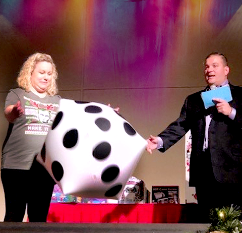 A contestant rolls a large inflatable die on stage during the Play it Forward charity team building game show event, while the host watches and holds game cards.