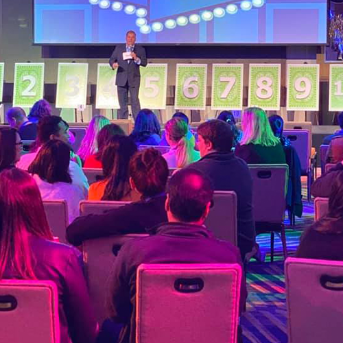 A host stands on stage in front of a row of numbered boards during the Play it Forward charity game show. The audience watches attentively as the host explains the next challenge in this interactive team building event, where all games and activities are designed to raise funds and donations for charity.