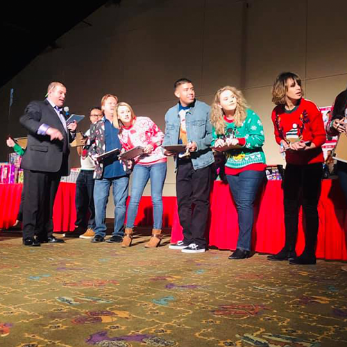 Contestants stand on stage during the Play it Forward charity game show, participating in a fun and engaging challenge led by the host. The contestants, wearing festive attire, eagerly await their next task in this team building event, where all proceeds and items are donated to charity.