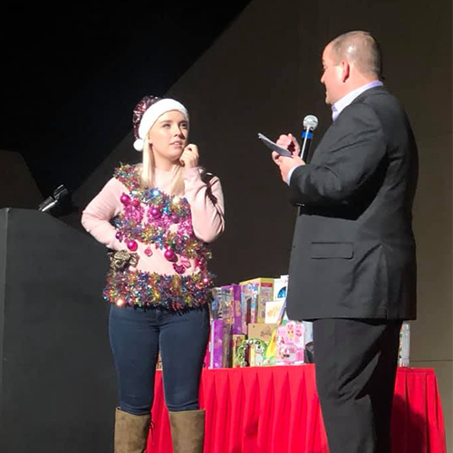 A contestant dressed in festive attire stands on stage with a host during the Play it Forward charitable team building game show. The host asks the contestant a question as they participate in one of the interactive challenges.