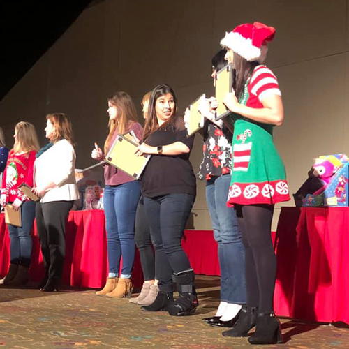 Participants stand in a row on stage during the Play it Forward charity game show, with some wearing festive holiday attire. Each holds a clipboard, preparing for a fun challenge as part of the team building event.