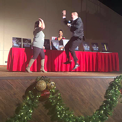 A contestant and host enthusiastically jump and high-five on stage during the Play it Forward charity game show. The event features exciting games like 'Stack the Deck,' with prizes and challenges, all aimed at raising donations for charity. The festive decorations add to the lively and energetic atmosphere of this team building activity.